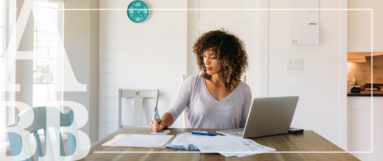 A woman sitting at a table with a laptop and paper documents, writing. 